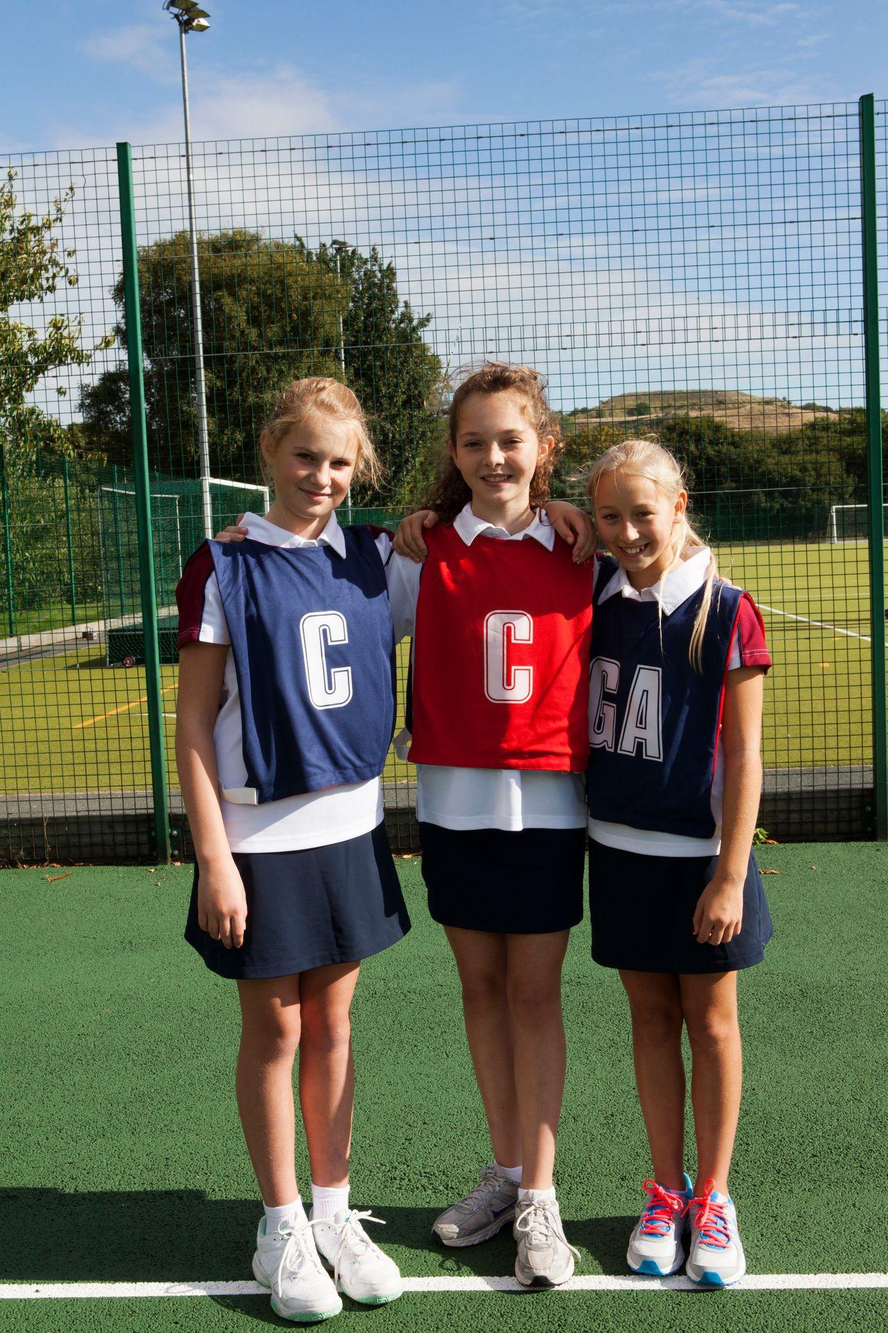 Three young girls in netball attire smiling on an outdoor court.